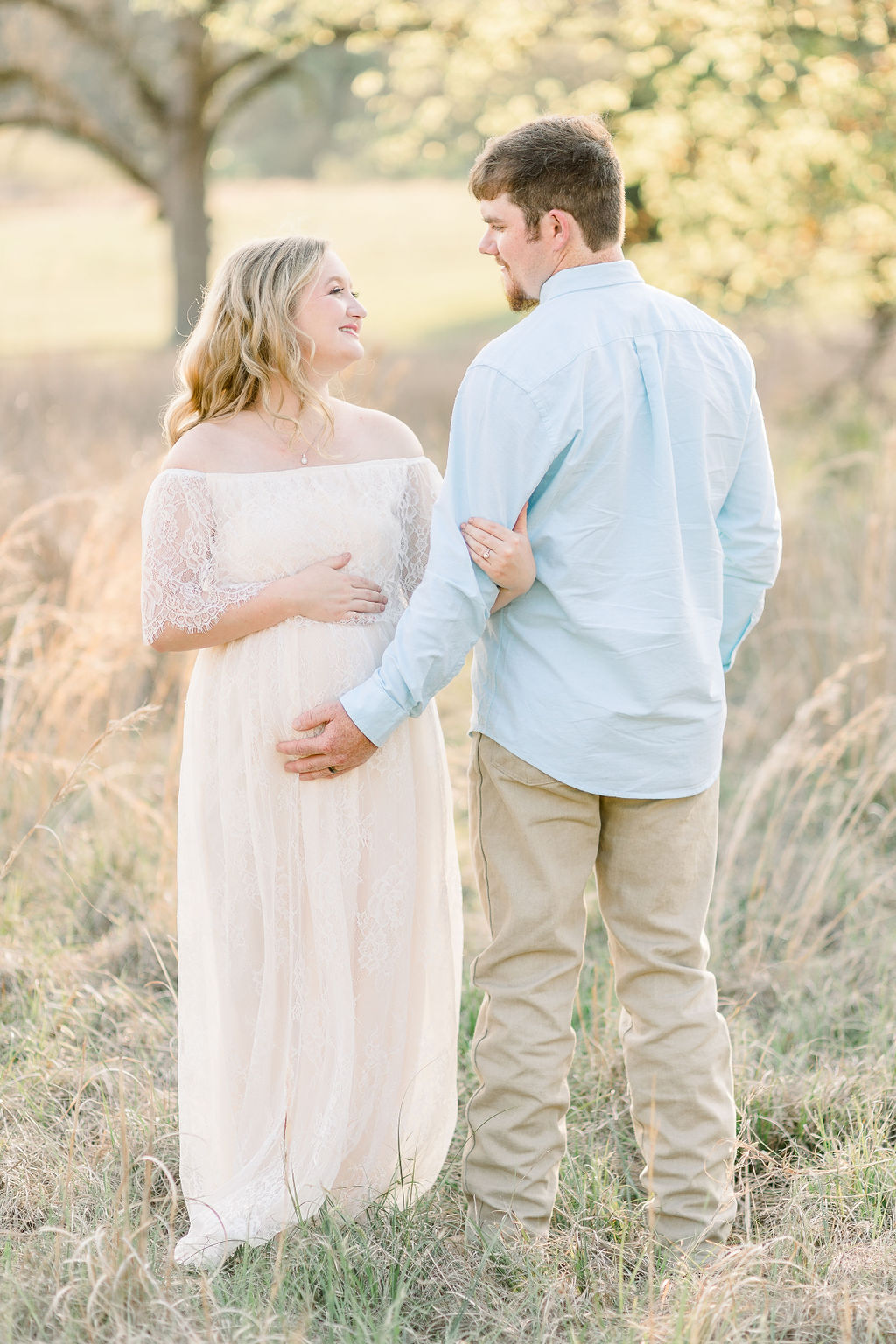 Mom in white lace dress holds onto dad's arm while they smile at each other and hold belly in green field. Image taken by Madison maternity photographer.