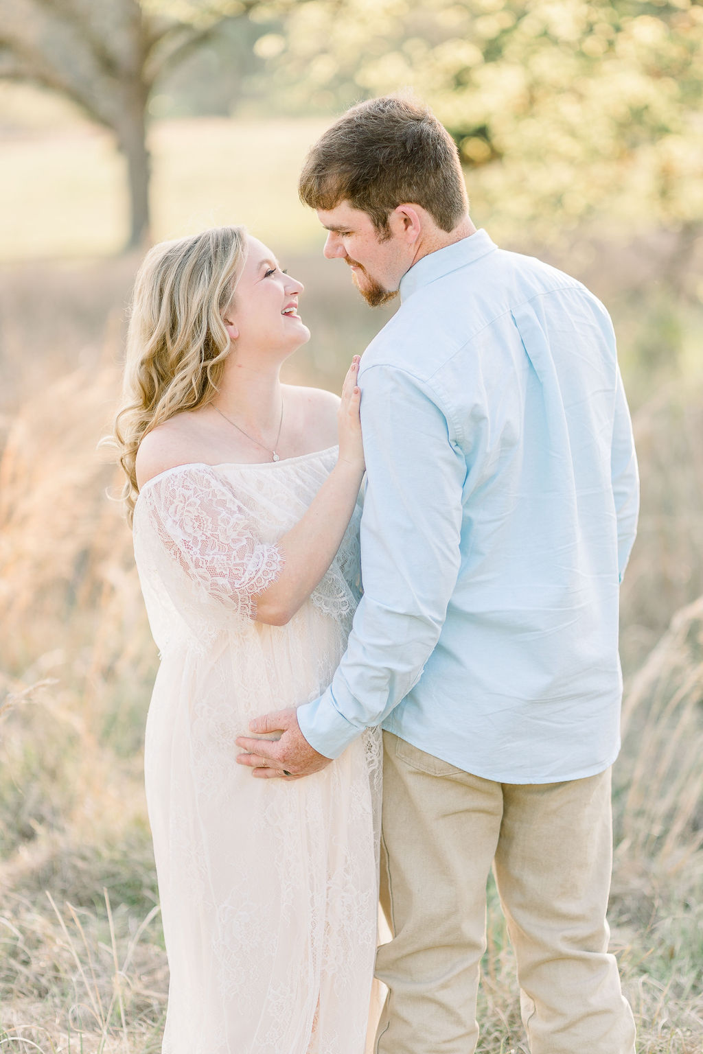 Mom in white lace dress holds onto dad while they smile at each other and hold belly in green field. Image taken by Madison maternity photographer.