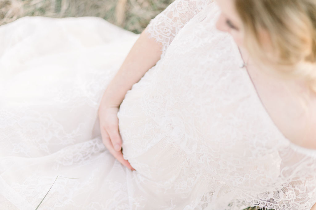 Close up image of maternity belly with mom wearing a white lace dress while sitting in a green field. Image by CJ and Olive Photography. 