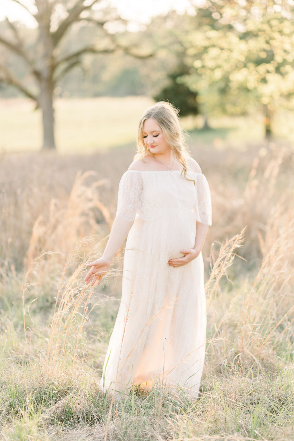 Mom to be in white lace maxi dress walking through a green grassy field at sunset at the Town of Livingston in Flora, Mississippi. Image taken by Madison maternity photographer.