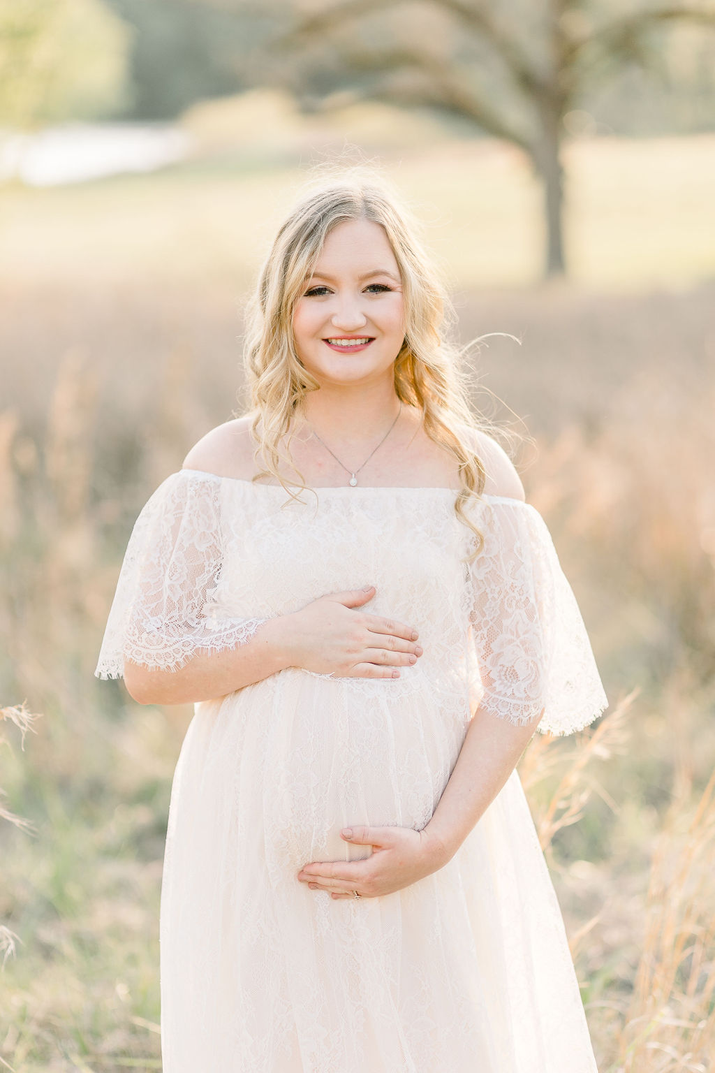 Mom to be smiles at camera while wearing white lace maxi dress in a green field at sunset. Image captured by CJ and Olive Photography, a Madison, Mississippi maternity photographer. 