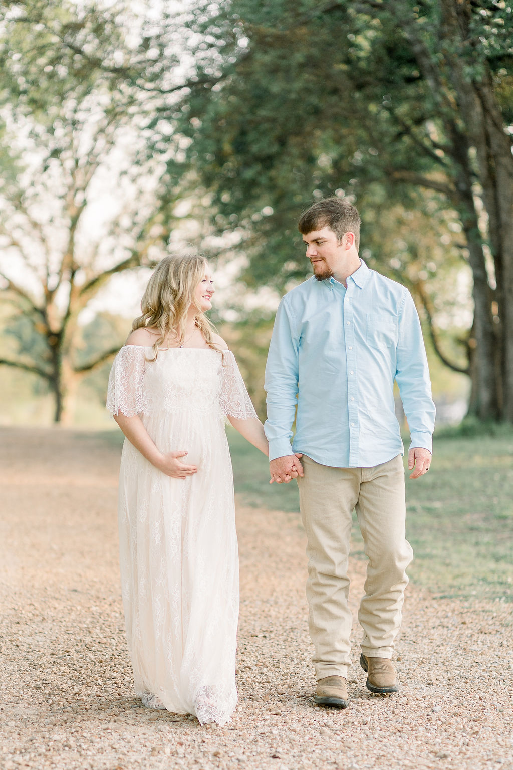 Mom in long white lace dress and dad  in light blue button up walk holding hands for maternity session in a green field. Image taken by Madison maternity photographer, CJ and Olive Photography.