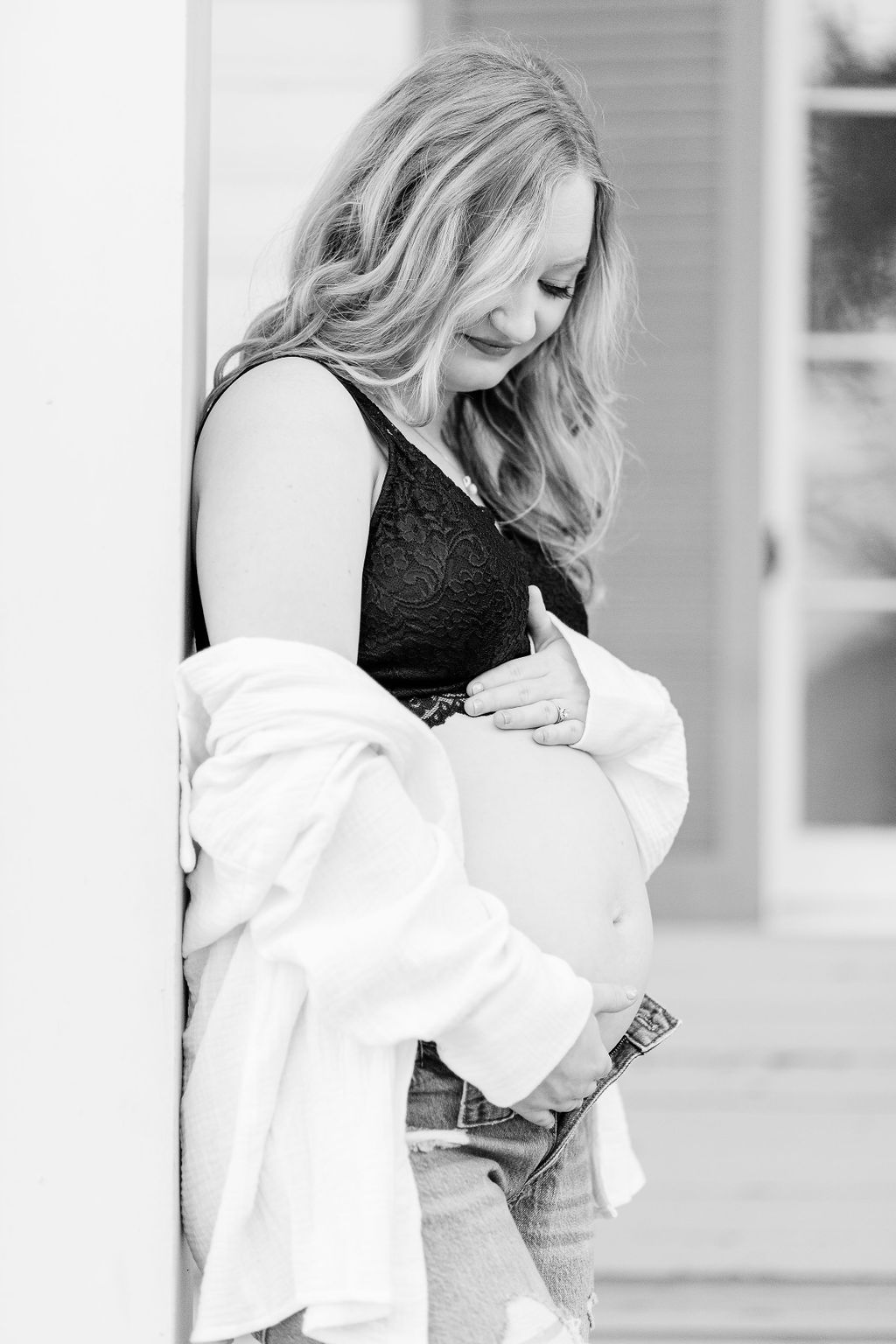 Mom to be looks down at belly while leaning on building post in white button up and jeans for maternity session. Image captured by CJ and Olive Photography. 