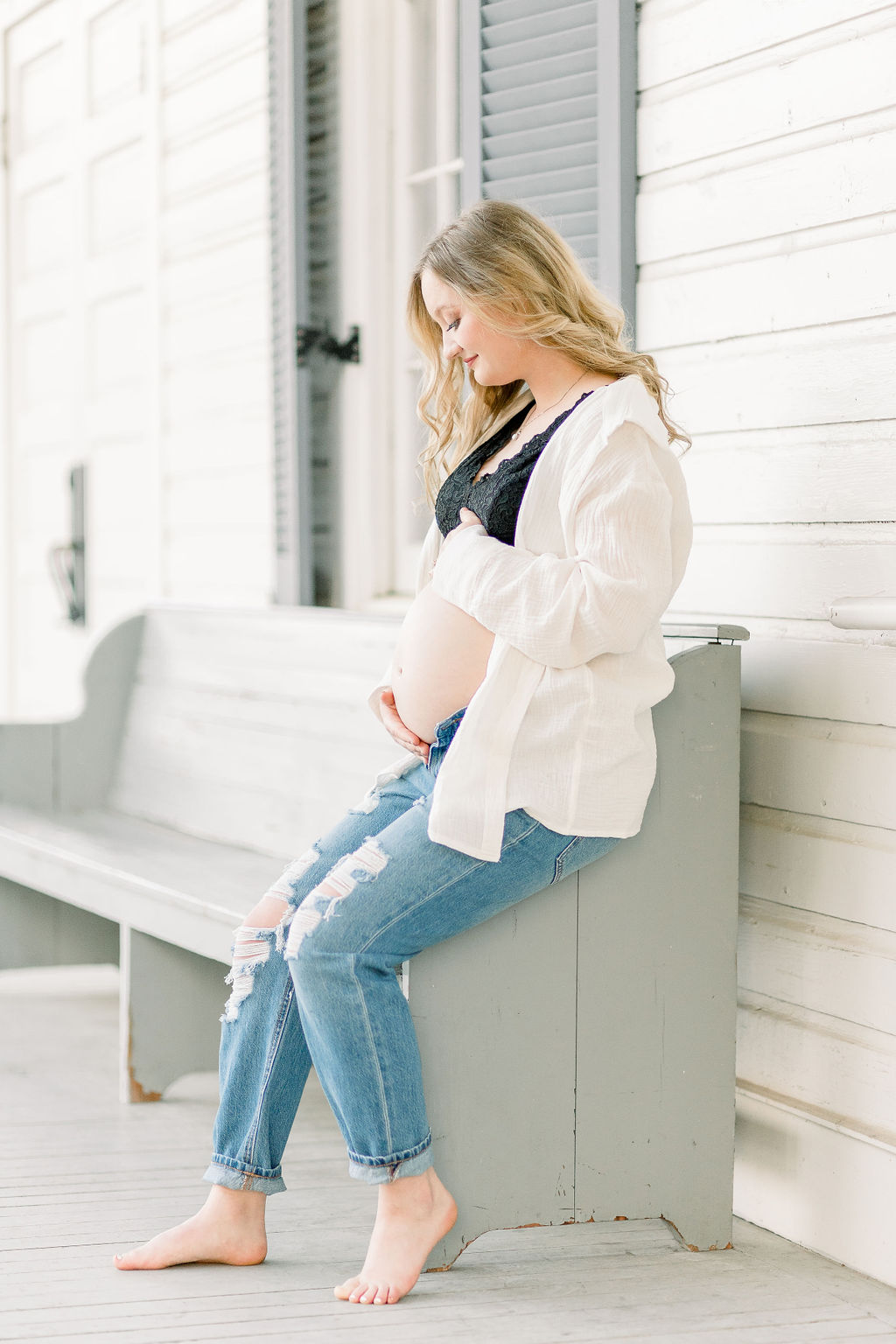 Mom to be looks down at belly while sitting on blue bench while wearing white button up shirt and blue jeans. Image taken by CJ and Olive Photography.