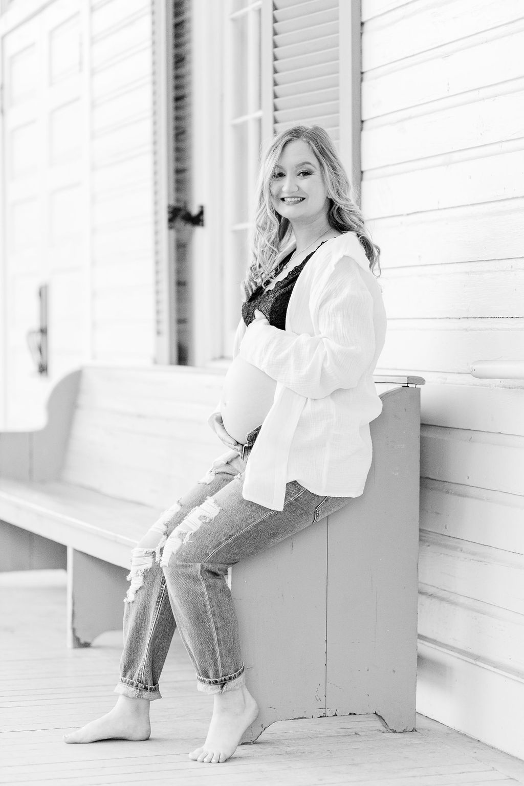 Mom to be smiles at camera holding belly while sitting on blue bench while wearing white button up shirt and blue jeans. Image taken by Madison Maternity Photographer. 