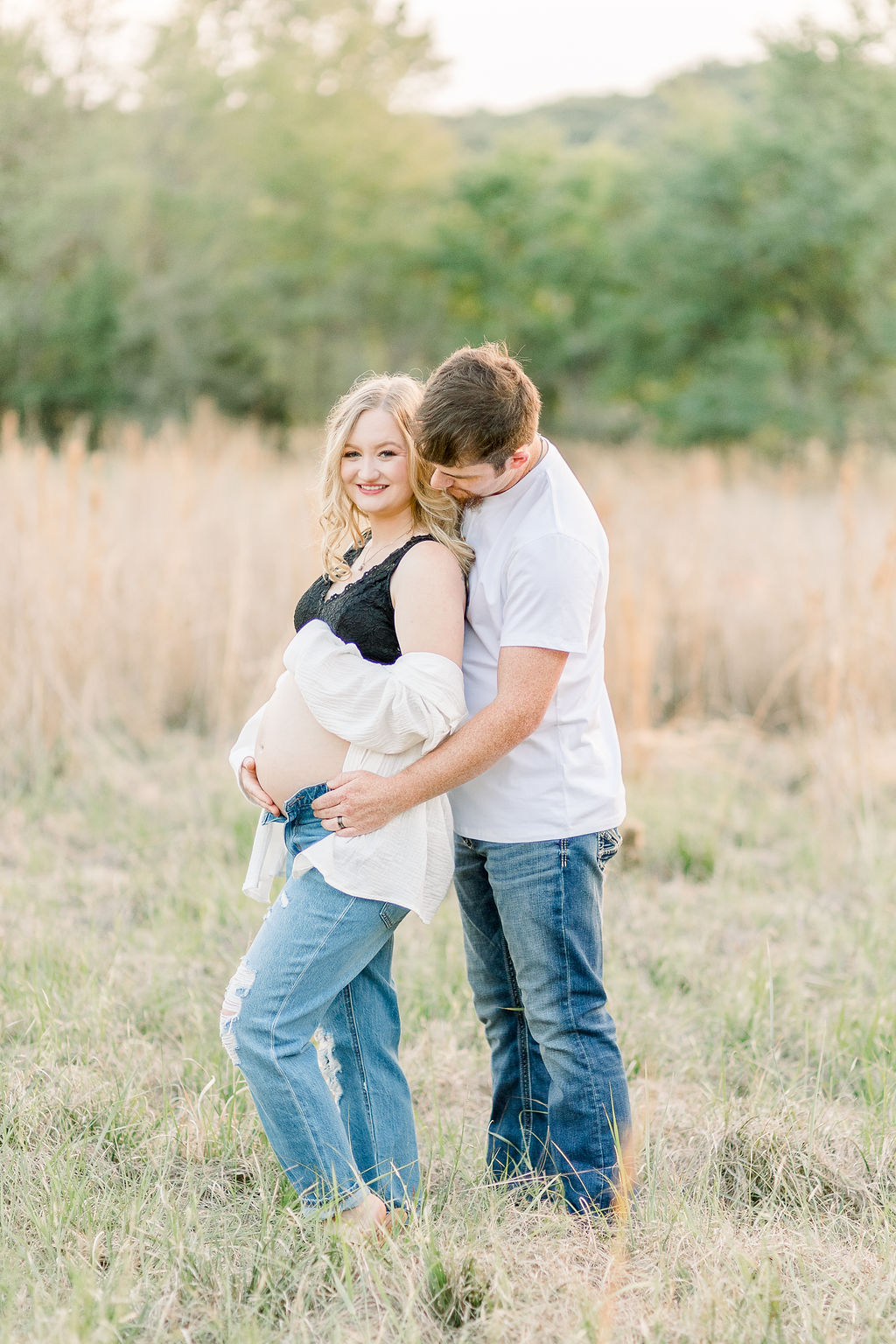 Mom and dad to be in green grassy field at sunset with dad behind mom and both hugging pregnant belly. Image captured by CJ and Olive Photography.