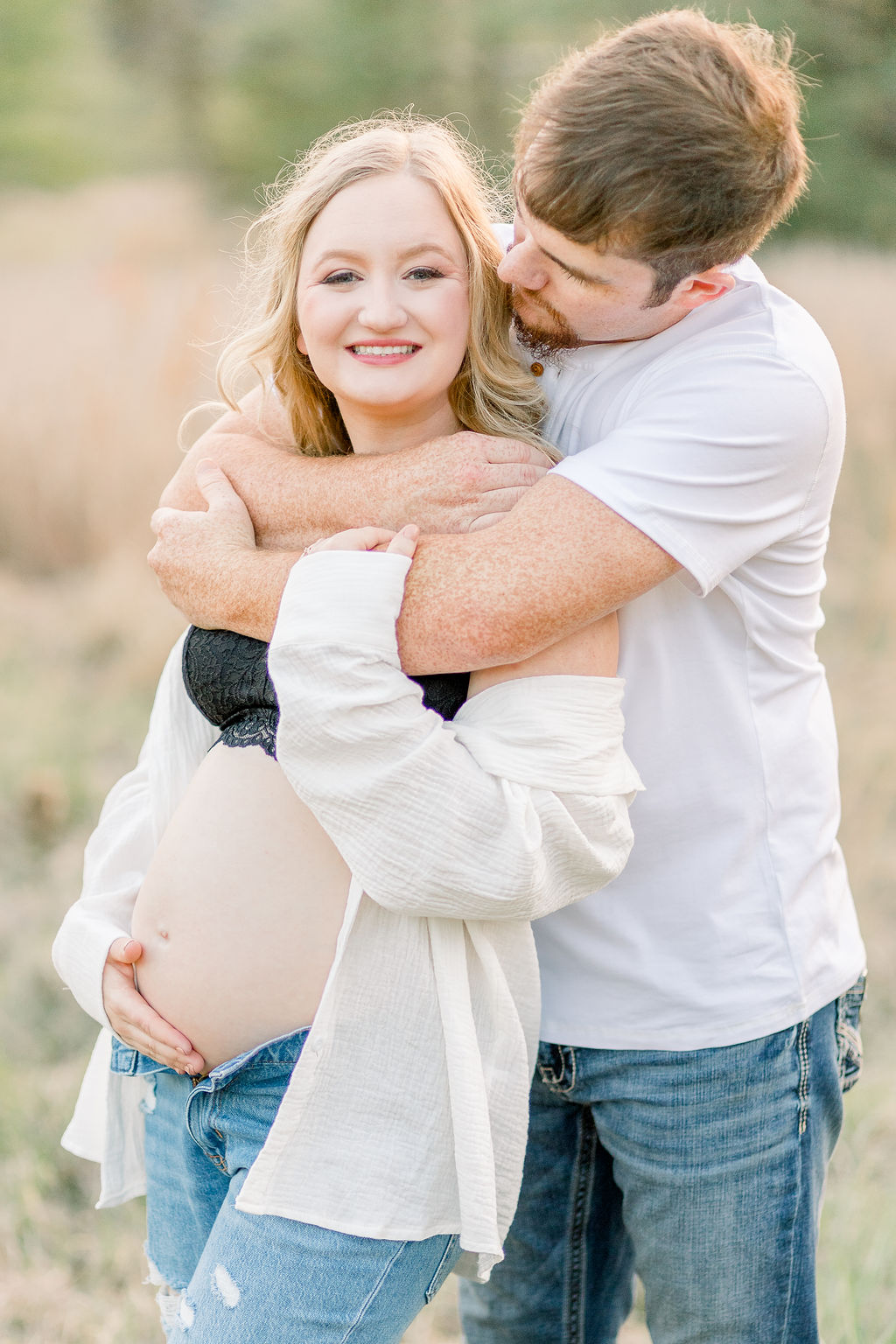 Mom and dad to be in green grassy field at sunset with dad behind mom and both hugging pregnant belly. Image captured by CJ and Olive Photography.