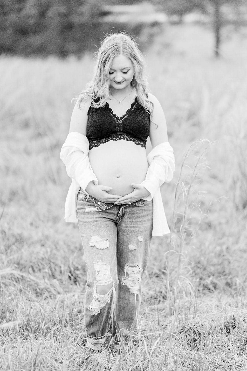 Mom to be shows off pregnant belly while standing in field  and smiling down at belly. Black and white image taken by CJ and Olive Photography.