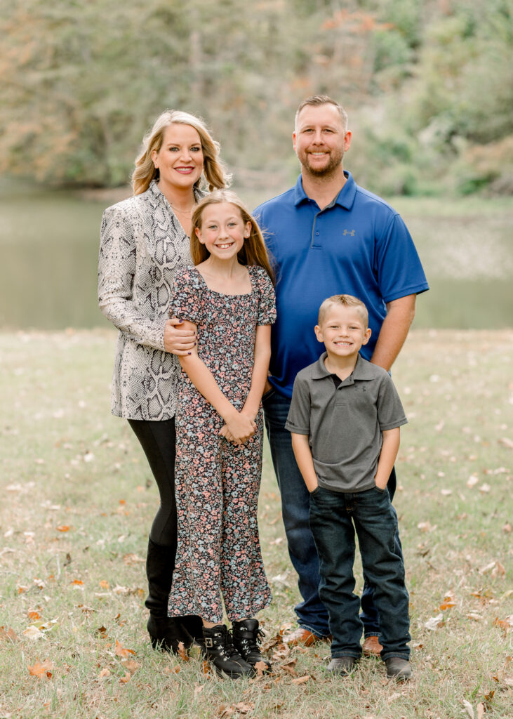 Family smiles together at camera while standing in front of pond for fall family photos by Mississippi Family Photographer. 