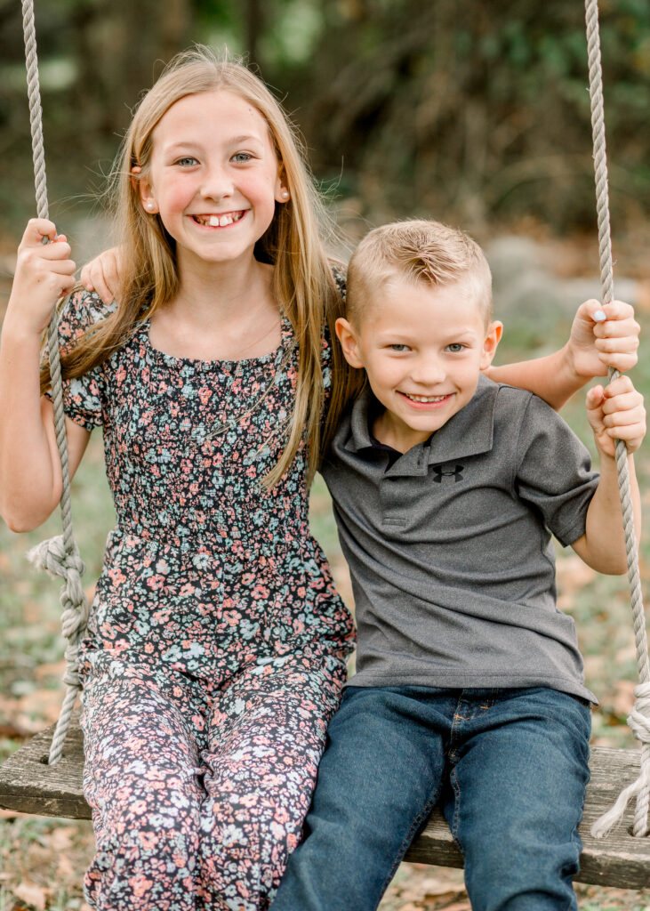 Brother and sister smile at camera while sitting on tree swing at The Resting Place in Flora, MS. Image captured by CJ and Olive Photography. 