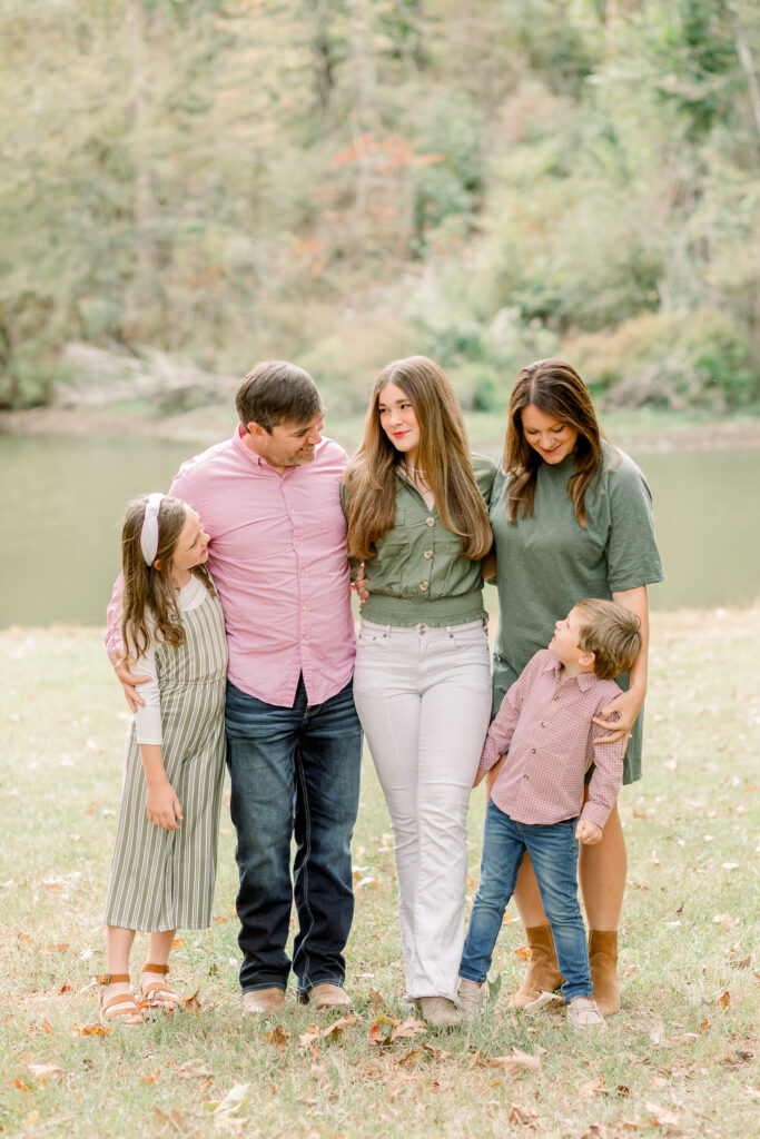 Family smiles together in front of pond at the Resting Place in Flora, MS  for fall family photos. Image captured by CJ and Olive Photography, a Mississippi family photographer. 