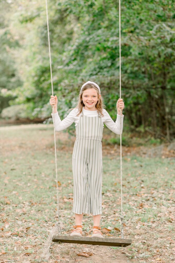 Little girl stands on tree swing and smiles at camera for fall family photos by Mississippi family photographer, CJ and Olive Photography. 