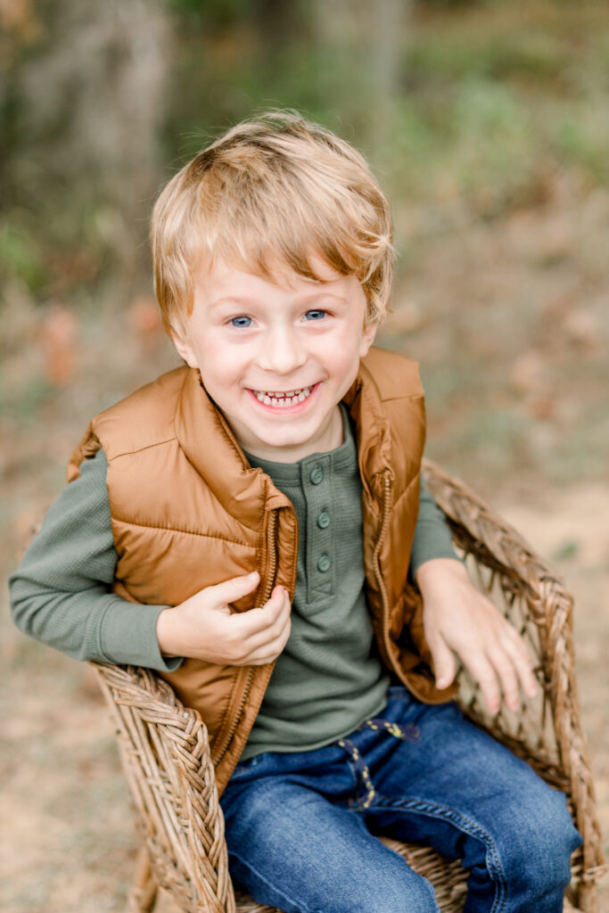 Little boy smiles at camera while sitting in a brown wicker chair. Image by CJ and Olive Photography, a Brandon, Mississippi photographer. 