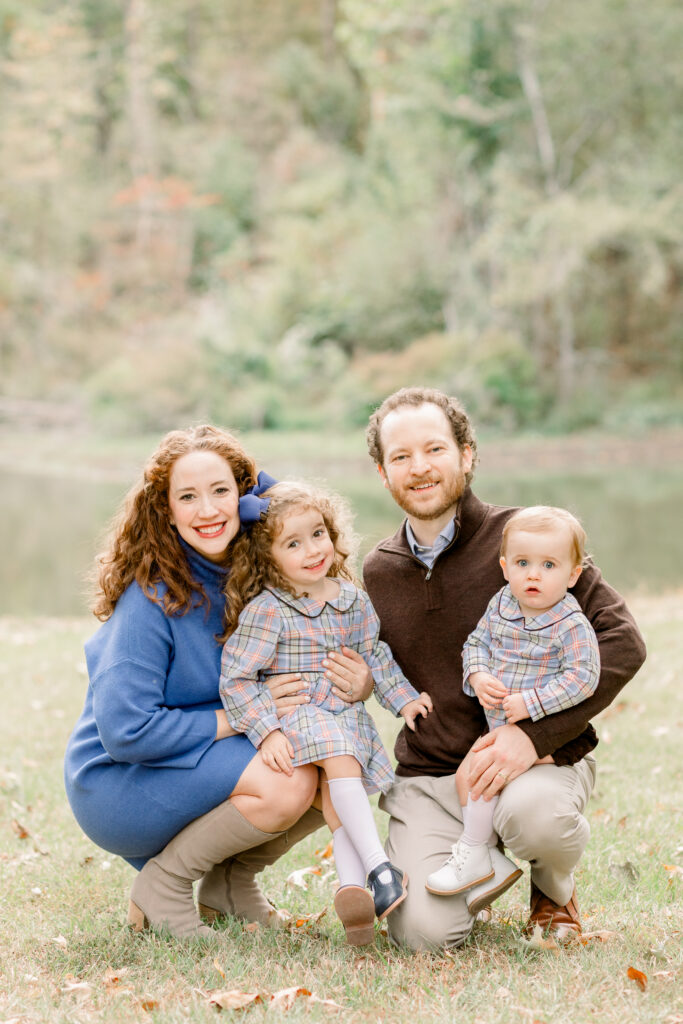 Family sits together in front of pond and smiles at the camera for fall family photos. Image captured by Madison family photographer, CJ and Olive Photography. 