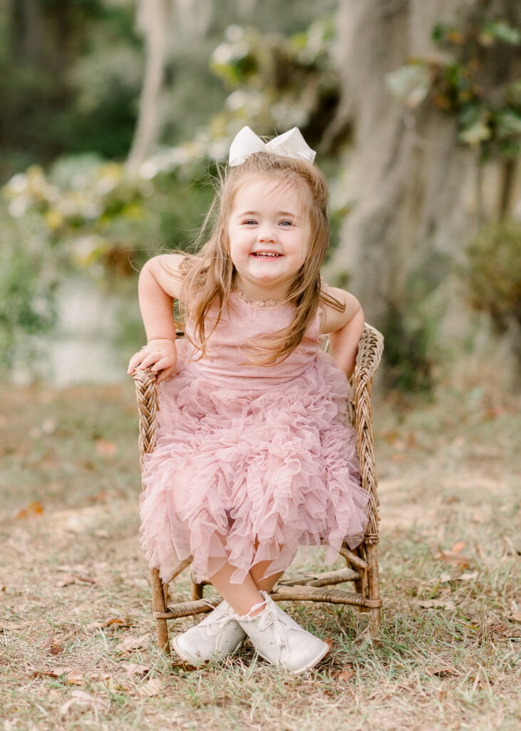 Little girl in pink dress smiles at camera while sitting in a small brown wicker chair for fall family session at the Resting place. Image captured by Brandon Family Photographer. 