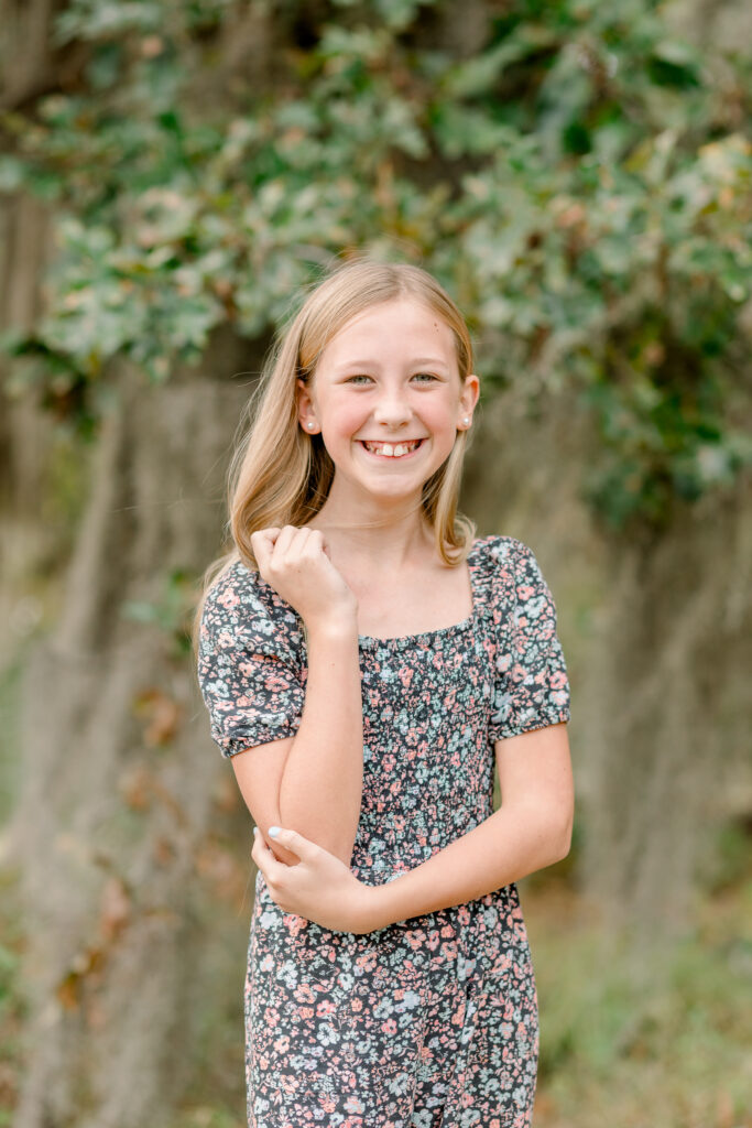 Tween girl in flower dress smiles at camera while playing with her hair for fall family photos by Mississippi Family Photographer.