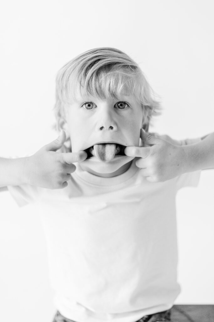 Black and white image of little boy sticking tongue out in white studio for personality portraits. Image captured by CJ and Olive Photography. 
