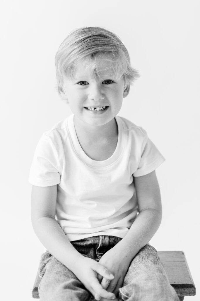 Black and white image of little boy smiling in white studio for personality portraits. Image captured by CJ and Olive Photography. 