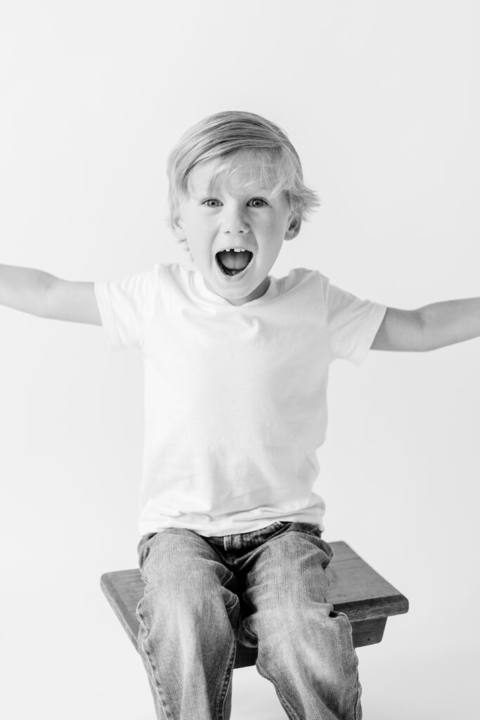 Black and white image taken by Brandon Family Photographer in white studio with little boy making an excited face with arms stretched out.  