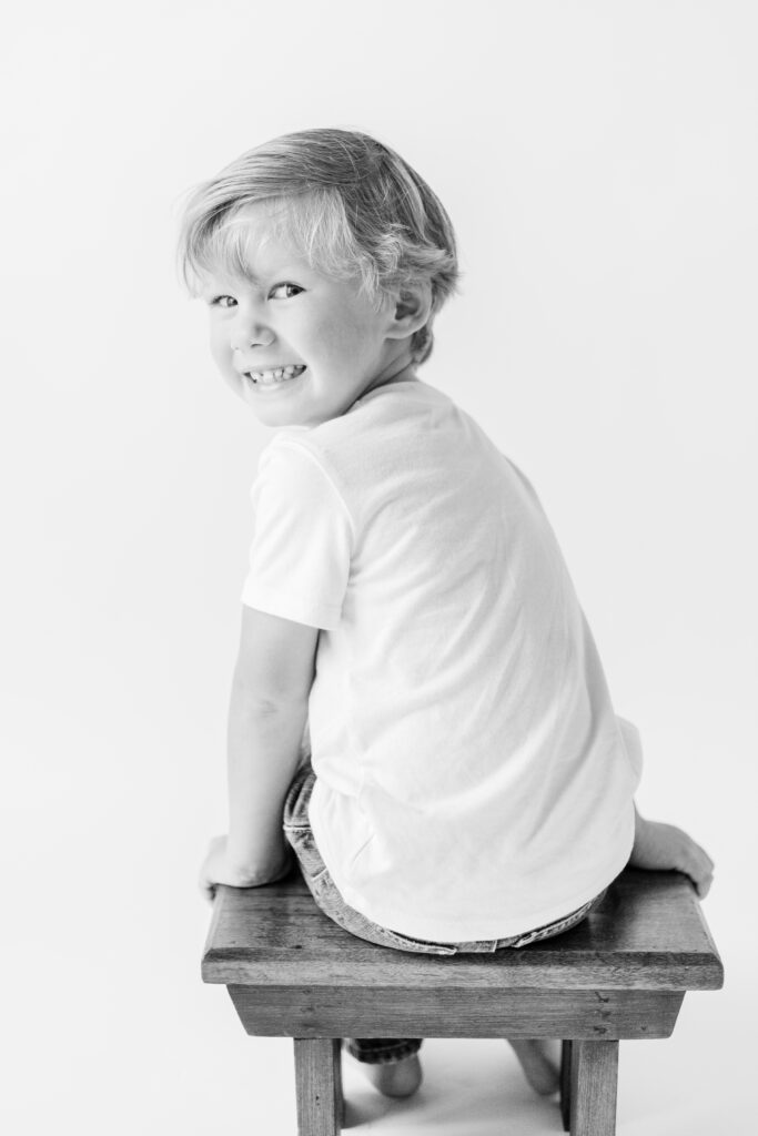 Black and white image of little boy smiling back in white studio for personality portraits. Image captured by CJ and Olive Photography. 